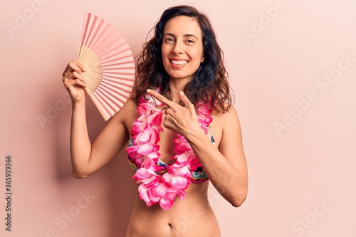 Young beautiful hispanic woman wearing bikini and hawaiian lei holding hand fan smiling happy pointing with hand and finger © Krakenimages.com