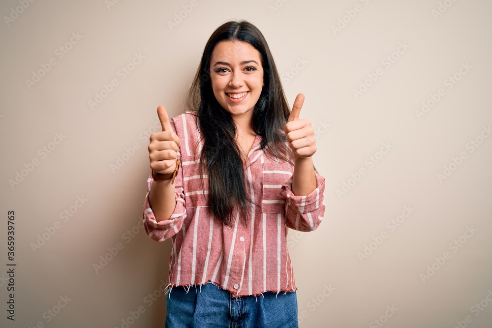 Young brunette woman wearing casual striped shirt over isolated background success sign doing positive gesture with hand, thumbs up smiling and happy. Cheerful expression and winner gesture. Young brunette woman wearing casual striped shirt over isolated background success sign doing positive gesture with hand, thumbs up smiling and happy. Cheerful expression and winner gesture.