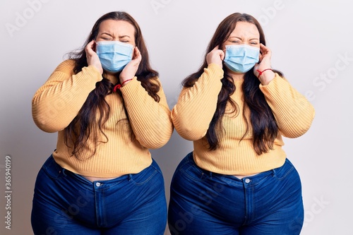 Young plus size twins wearing medical mask covering ears with fingers with annoyed expression for the noise of loud music. deaf concept. photo
