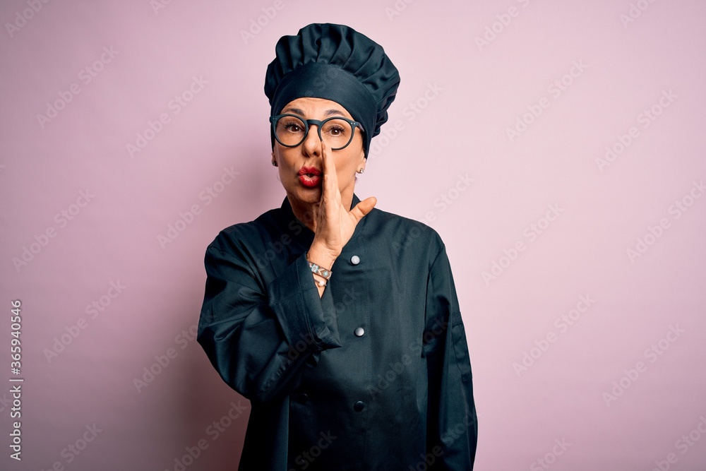 Middle age brunette chef woman wearing cooker uniform and hat over isolated pink background hand on mouth telling secret rumor, whispering malicious talk conversation