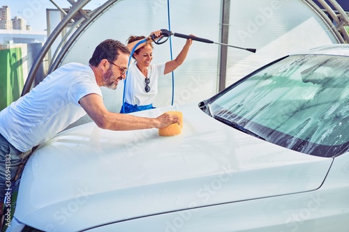 Middle age beautiful couple wearing casual clothes and smiling happy. Standing with smile on face washing car using water pistol and sponge.