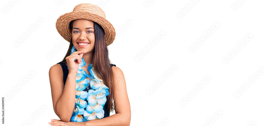 Young beautiful latin girl wearing hawaiian lei and summer hat looking confident at the camera with smile with crossed arms and hand raised on chin. thinking positive. Young beautiful latin girl wearing hawaiian lei and summer hat looking confident at the camera with smile with crossed arms and hand raised on chin. thinking positive.