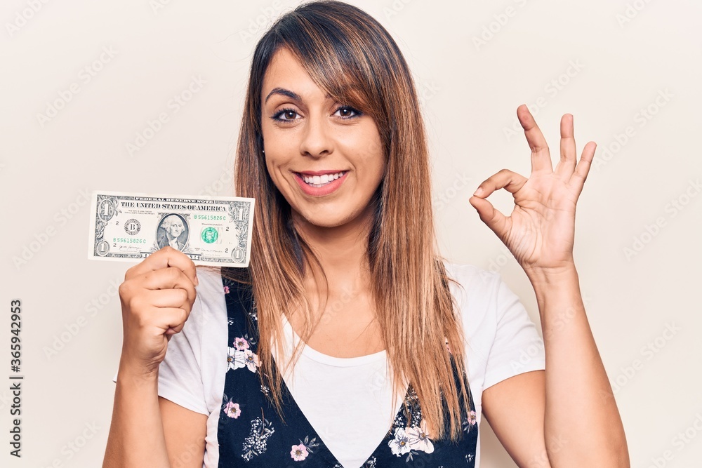 Young beautiful woman holding one dollar doing ok sign with fingers, smiling friendly gesturing excellent symbol Young beautiful woman holding one dollar doing ok sign with fingers, smiling friendly gesturing excellent symbol