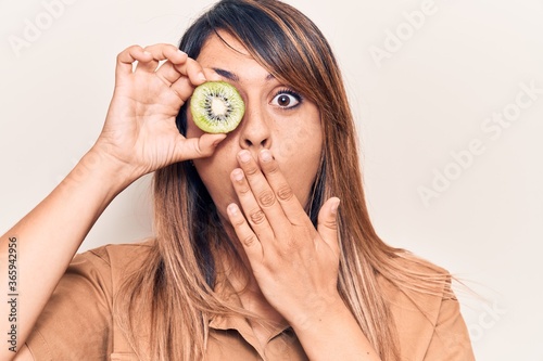 Young beautiful woman holding kiwi over eye covering mouth with hand, shocked and afraid for mistake. surprised expression