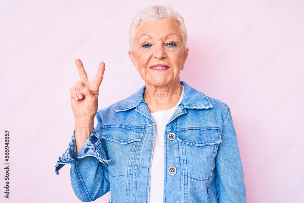 Senior beautiful woman with blue eyes and grey hair wearing casual denim jacket smiling with happy face winking at the camera doing victory sign. number two. Senior beautiful woman with blue eyes and grey hair wearing casual denim jacket smiling with happy face winking at the camera doing victory sign. number two.