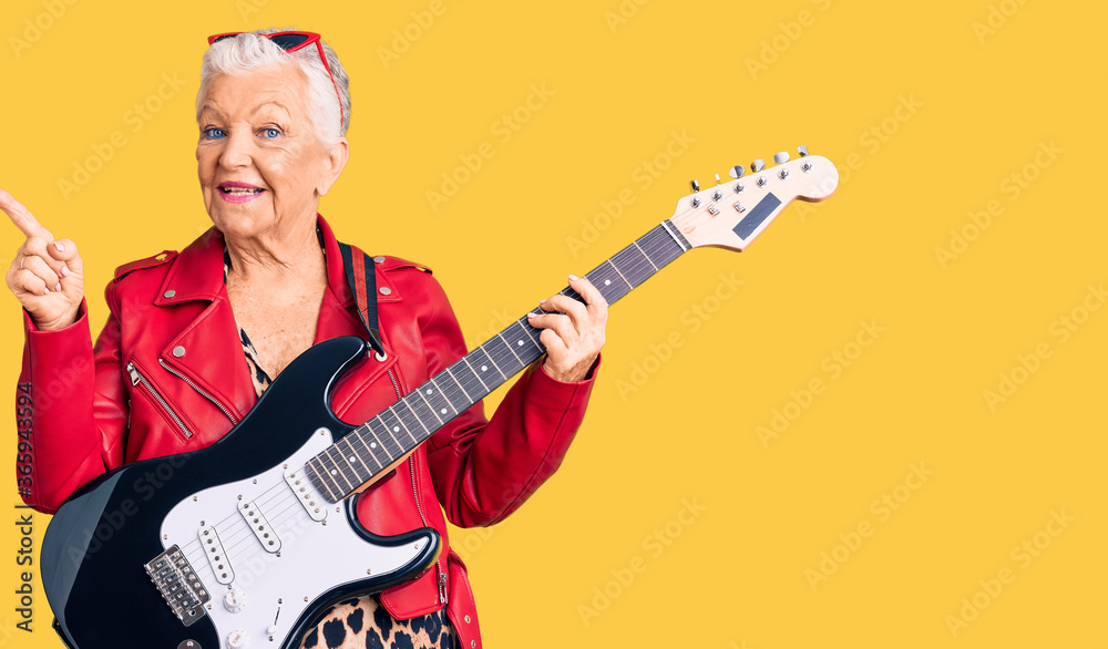 Senior beautiful woman with blue eyes and grey hair wearing a modern look playing electric guitar with a big smile on face, pointing with hand finger to the side looking at the camera.
