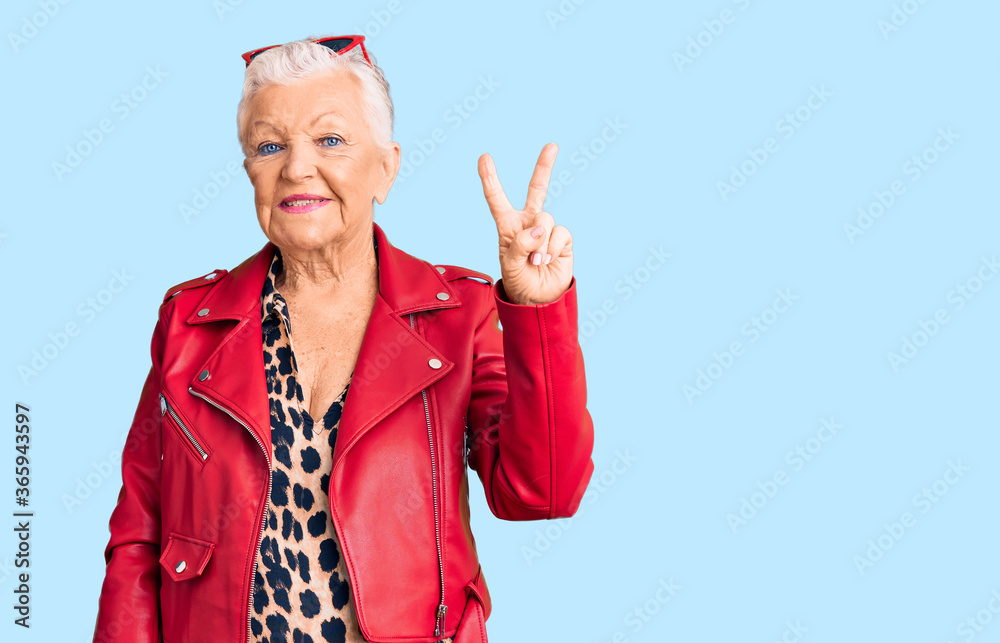 Senior beautiful woman with blue eyes and grey hair wearing a modern style with a red leather jacket smiling with happy face winking at the camera doing victory sign. number two. Senior beautiful woman with blue eyes and grey hair wearing a modern style with a red leather jacket smiling with happy face winking at the camera doing victory sign. number two.