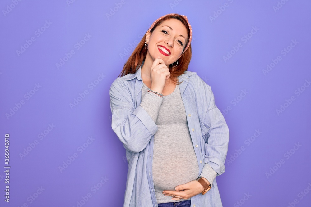 Young beautiful redhead pregnant woman expecting baby over isolated purple background looking confident at the camera with smile with crossed arms and hand raised on chin. Thinking positive. Young beautiful redhead pregnant woman expecting baby over isolated purple background looking confident at the camera with smile with crossed arms and hand raised on chin. Thinking positive.