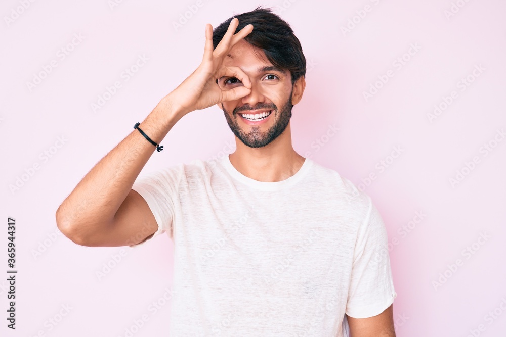 Handsome hispanic man wearing casual white tshirt smiling happy doing ok sign with hand on eye looking through fingers Handsome hispanic man wearing casual white tshirt smiling happy doing ok sign with hand on eye looking through fingers