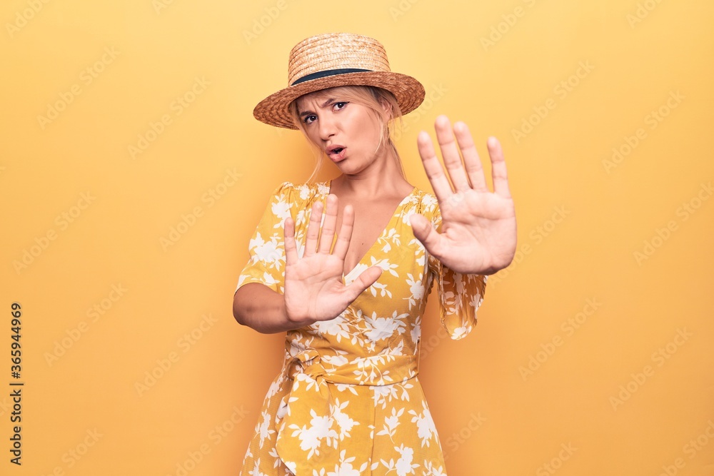 Beautiful blonde woman on vacation wearing summer hat and dress over yellow background Moving away hands palms showing refusal and denial with afraid and disgusting expression. Stop and forbidden.