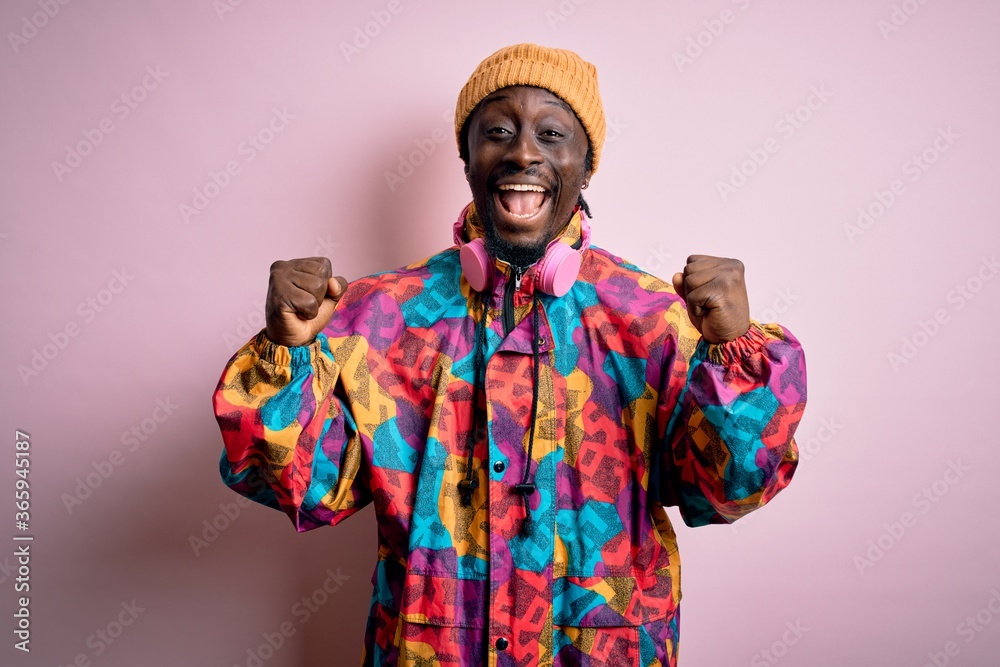 Young handsome african american man wearing colorful coat and cap over pink background celebrating surprised and amazed for success with arms raised and open eyes. Winner concept.