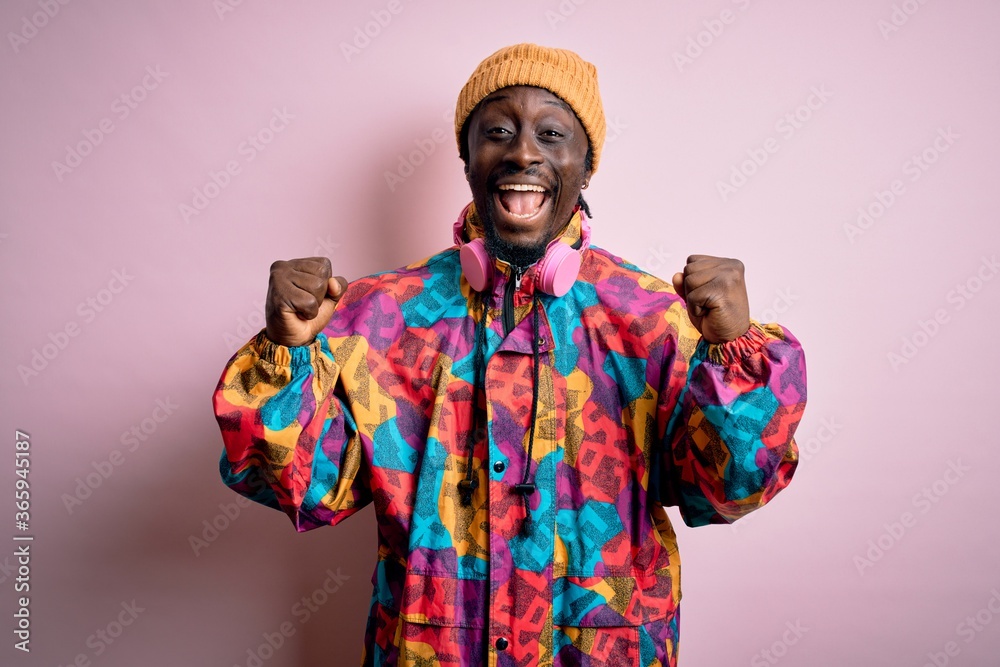 Obraz premium Young handsome african american man wearing colorful coat and cap over pink background celebrating surprised and amazed for success with arms raised and open eyes. Winner concept.