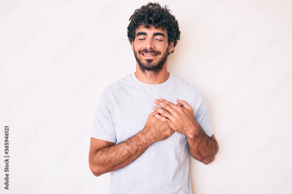 Handsome young man with curly hair and bear wearing casual tshirt smiling with hands on chest with closed eyes and grateful gesture on face. health concept. Handsome young man with curly hair and bear wearing casual tshirt smiling with hands on chest with closed eyes and grateful gesture on face. health concept.