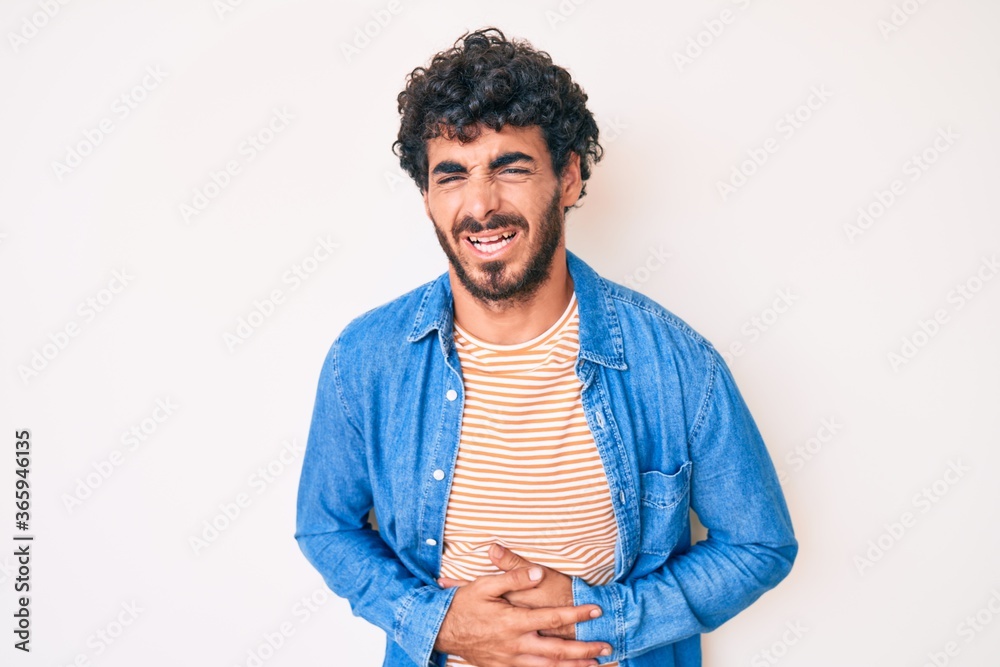Handsome young man with curly hair and bear wearing casual denim jacket with hand on stomach because nausea, painful disease feeling unwell. ache concept. Handsome young man with curly hair and bear wearing casual denim jacket with hand on stomach because nausea, painful disease feeling unwell. ache concept.