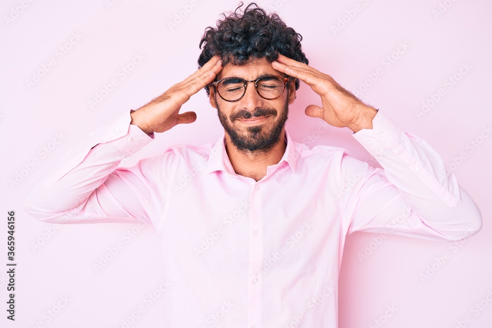 Handsome young man with curly hair and bear wearing business shirt and glasses suffering from headache desperate and stressed because pain and migraine. hands on head. Handsome young man with curly hair and bear wearing business shirt and glasses suffering from headache desperate and stressed because pain and migraine. hands on head.