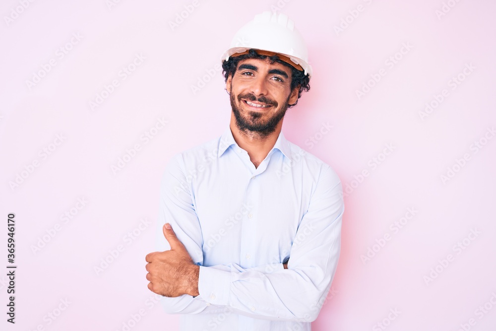 Handsome young man with curly hair and bear wearing architect hardhat happy face smiling with crossed arms looking at the camera. positive person.