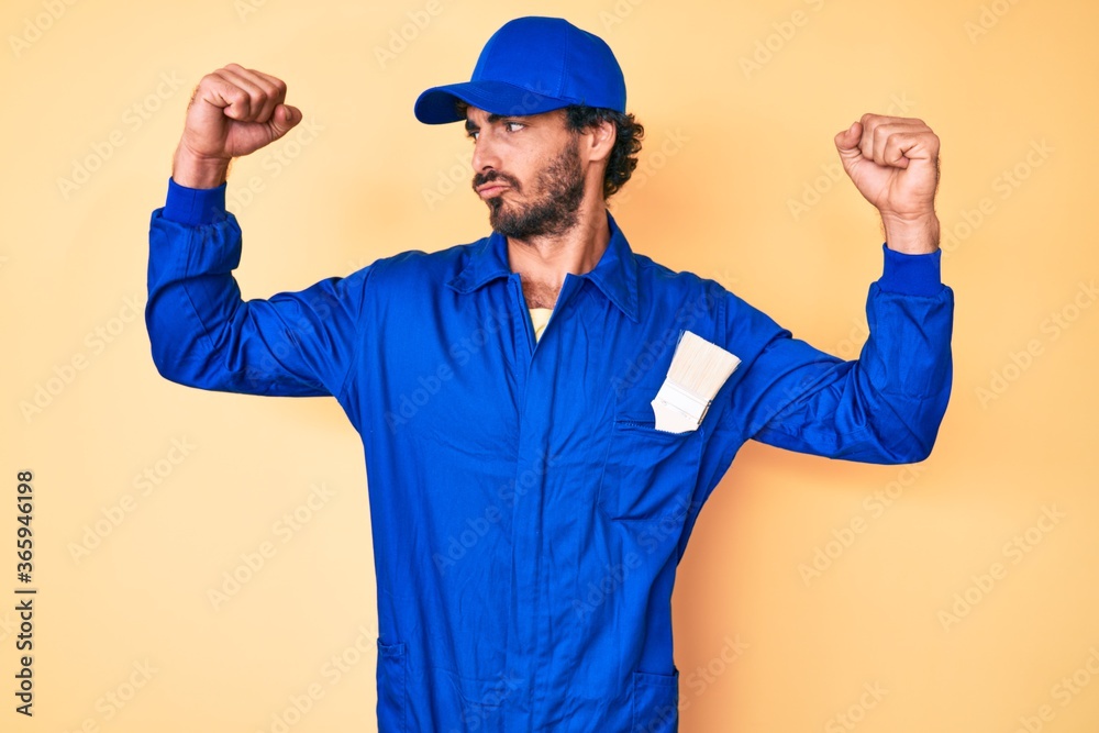 Handsome young man with curly hair and bear wearing builder jumpsuit uniform showing arms muscles smiling proud. fitness concept.