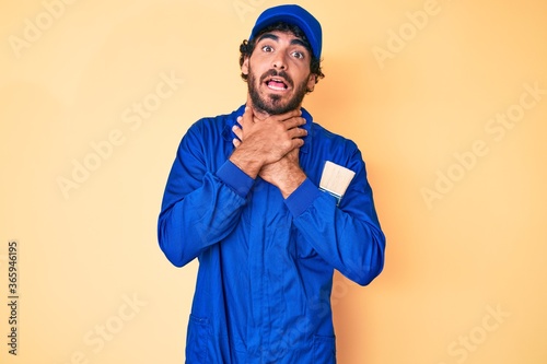 Handsome young man with curly hair and bear wearing builder jumpsuit uniform shouting and suffocate because painful strangle. health problem. asphyxiate and suicide concept.