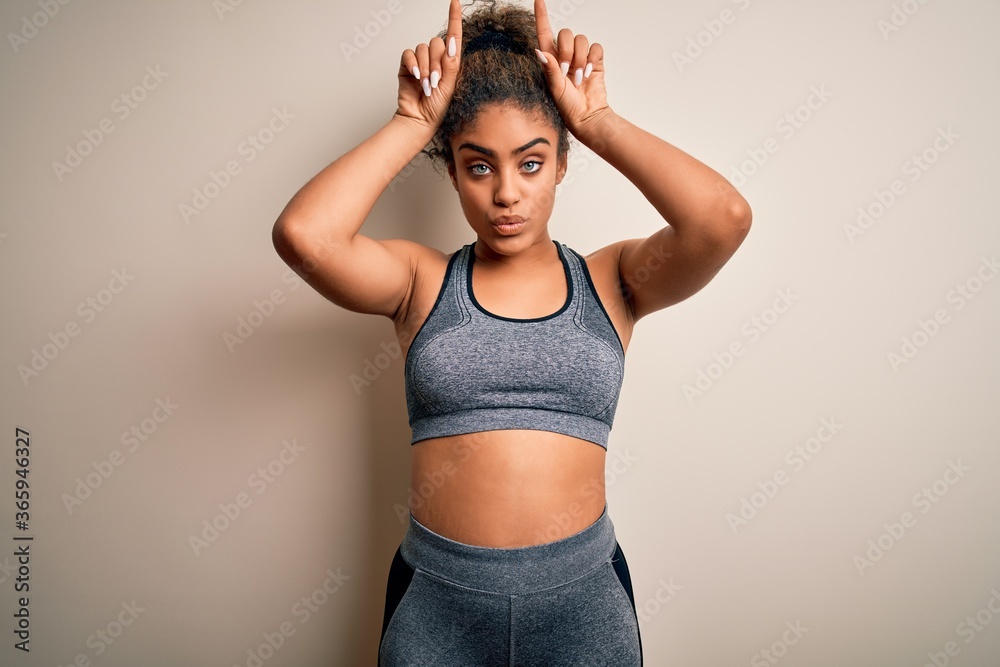 Young african american sportswoman doing sport wearing sportswear over white background doing funny gesture with finger over head as bull horns Young african american sportswoman doing sport wearing sportswear over white background doing funny gesture with finger over head as bull horns