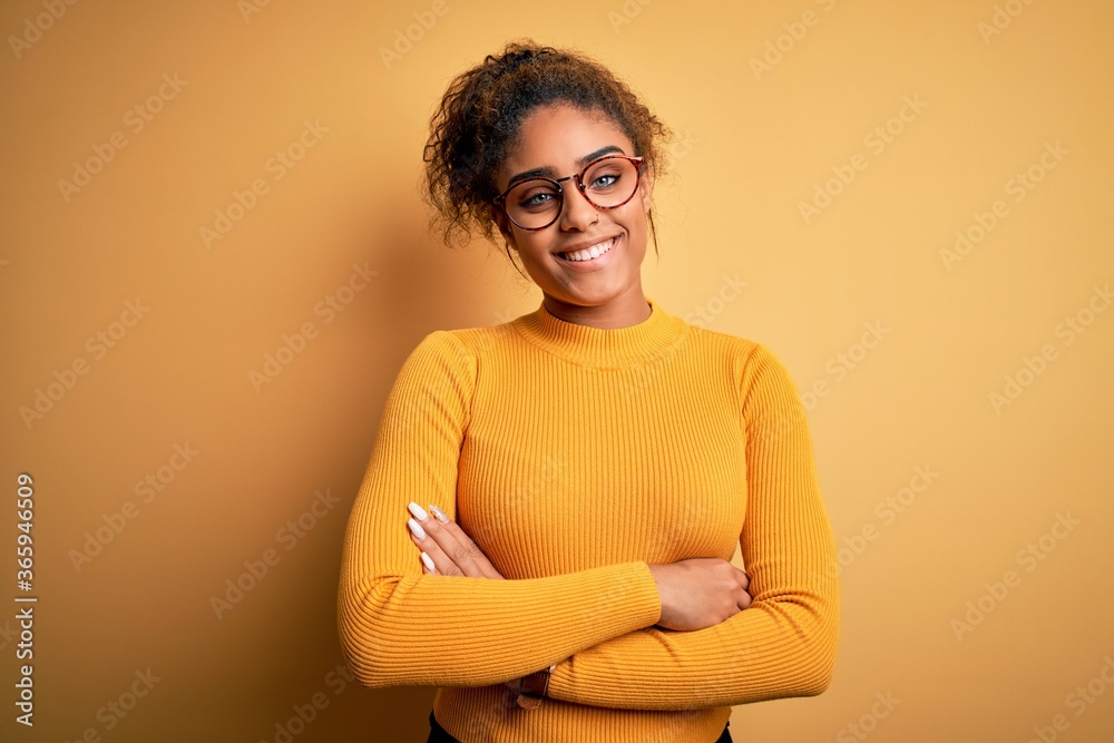 Young beautiful african american girl wearing sweater and glasses over yellow background happy face smiling with crossed arms looking at the camera. Positive person. Young beautiful african american girl wearing sweater and glasses over yellow background happy face smiling with crossed arms looking at the camera. Positive person.