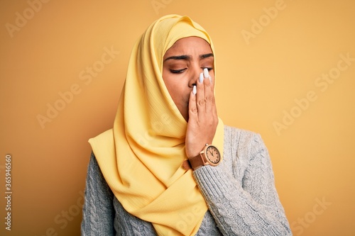 Young beautiful african american girl wearing muslim hijab over isolated yellow background bored yawning tired covering mouth with hand. Restless and sleepiness. © Krakenimages.com