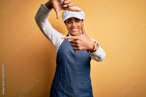 Young african american afro baker woman wearing apron and cap over yellow background smiling making frame with hands and fingers with happy face. Creativity and photography concept.