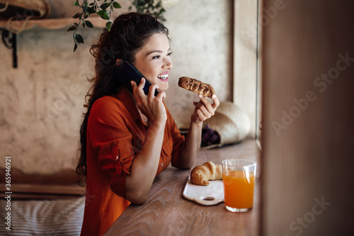 Beautiful young woman talking on phone while eating delicious rolls in bakery.