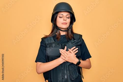 Obraz na płótnie Young beautiful brunette soldier woman wearing bulletproof and security helmet smiling with hands on chest with closed eyes and grateful gesture on face