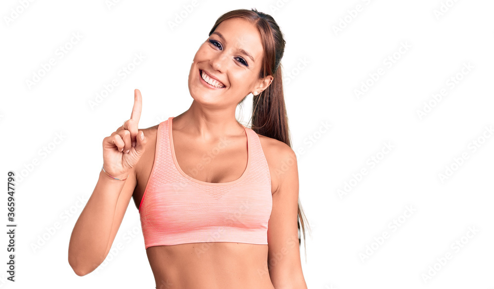 Young beautiful hispanic woman wearing sportswear showing and pointing up with finger number one while smiling confident and happy. Young beautiful hispanic woman wearing sportswear showing and pointing up with finger number one while smiling confident and happy.