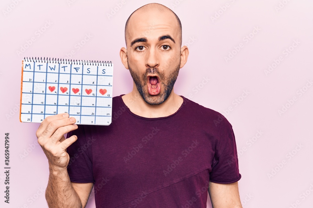 Young handsome bald man holding heart calendar scared and amazed with open mouth for surprise, disbelief face Young handsome bald man holding heart calendar scared and amazed with open mouth for surprise, disbelief face