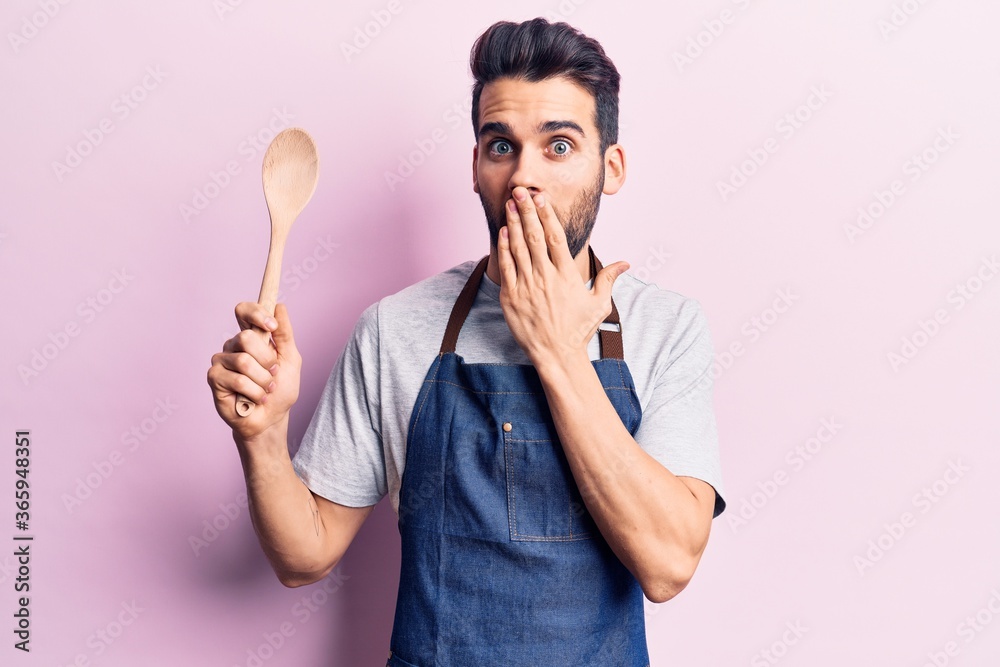Young handsome man with beard wearing apron holding wooden spoon covering mouth with hand, shocked and afraid for mistake. surprised expression Young handsome man with beard wearing apron holding wooden spoon covering mouth with hand, shocked and afraid for mistake. surprised expression