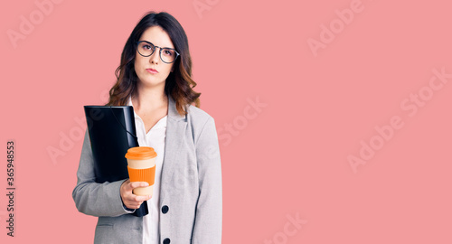 Beautiful young brunette woman holding business folder and drinking coffee thinking attitude and sober expression looking self confident