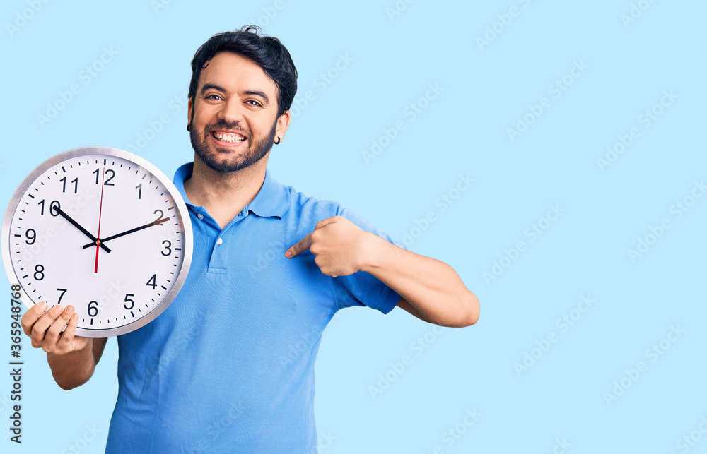 Young hispanic man holding big clock pointing finger to one self smiling happy and proud Young hispanic man holding big clock pointing finger to one self smiling happy and proud