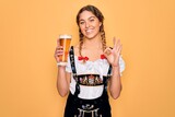 Beautiful blonde german woman with blue eyes wearing octoberfest dress drinking glass of beer doing ok sign with fingers, excellent symbol