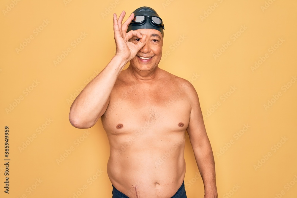 Middle age senior grey-haired swimmer man wearing swimsuit, cap and goggles doing ok gesture with hand smiling, eye looking through fingers with happy face.