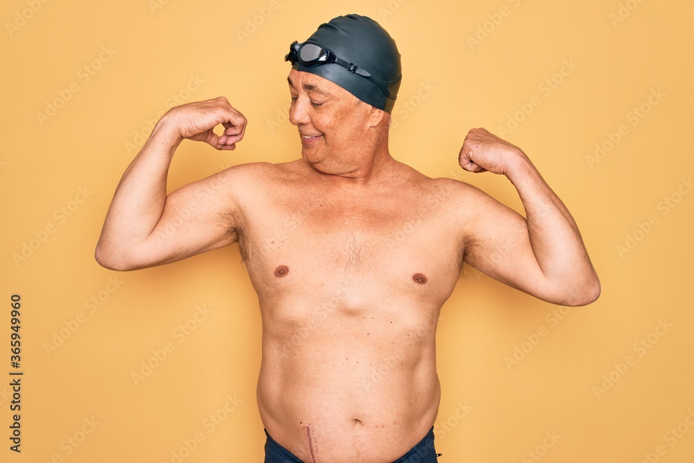 Middle age senior grey-haired swimmer man wearing swimsuit, cap and goggles showing arms muscles smiling proud. Fitness concept.