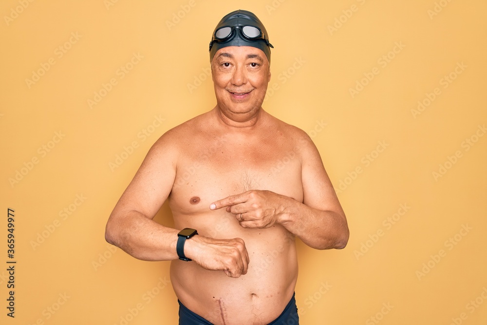 Middle age senior grey-haired swimmer man wearing swimsuit, cap and goggles In hurry pointing to watch time, impatience, upset and angry for deadline delay