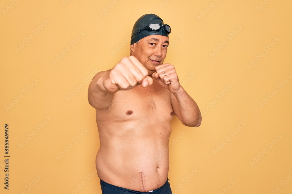 Middle age senior grey-haired swimmer man wearing swimsuit, cap and goggles Punching fist to fight, aggressive and angry attack, threat and violence