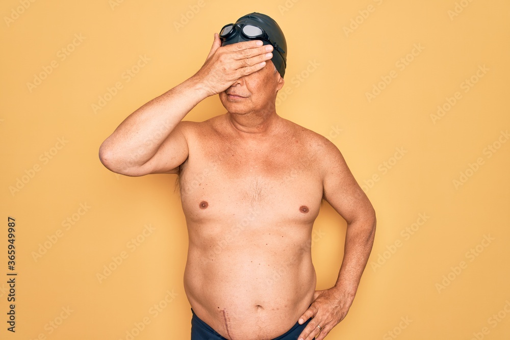Middle age senior grey-haired swimmer man wearing swimsuit, cap and goggles covering eyes with hand, looking serious and sad. Sightless, hiding and rejection concept