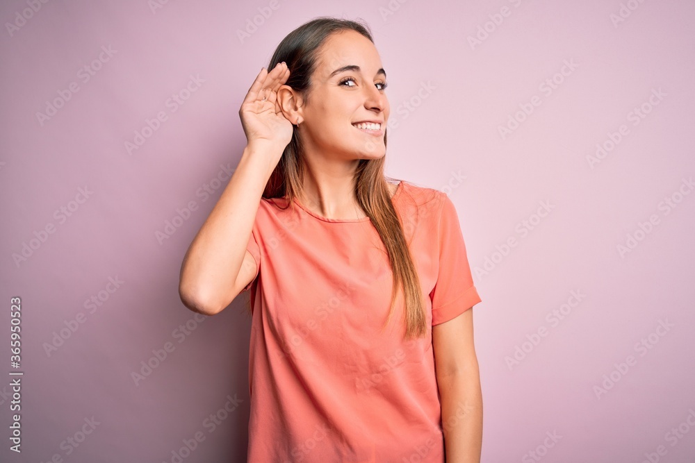 Young beautiful woman wearing casual t-shirt standing over isolated pink background smiling with hand over ear listening an hearing to rumor or gossip. Deafness concept. Young beautiful woman wearing casual t-shirt standing over isolated pink background smiling with hand over ear listening an hearing to rumor or gossip. Deafness concept.