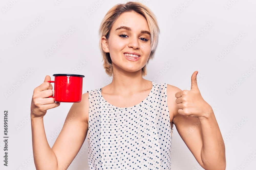 Young beautiful blonde woman holding coffee smiling happy and positive, thumb up doing excellent and approval sign Young beautiful blonde woman holding coffee smiling happy and positive, thumb up doing excellent and approval sign