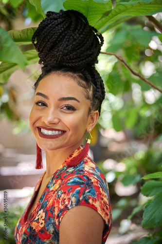 Stunning young biracial woman stands outside wearing floral print dress with hair braided and up - near plants