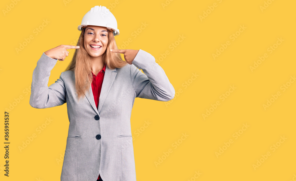 Beautiful young woman wearing architect hardhat smiling cheerful showing and pointing with fingers teeth and mouth. dental health concept.