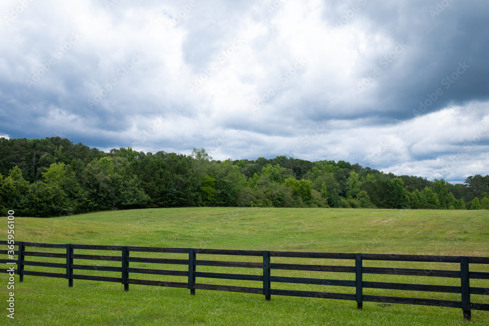Storm clouds over a brilliant green field, black rail fence in front with treeline on the horizon, horizontal aspect