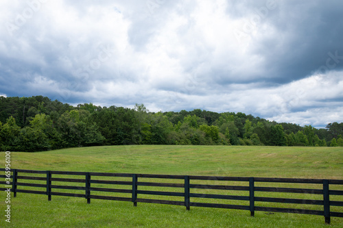 Storm clouds over a brilliant green field, black rail fence in front with treeline on the horizon, horizontal aspect