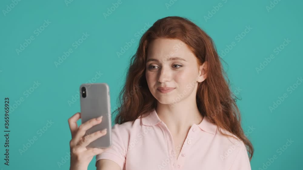 Beautiful smiling redhead girl excitedly talking on smartphone by video call over colorful background