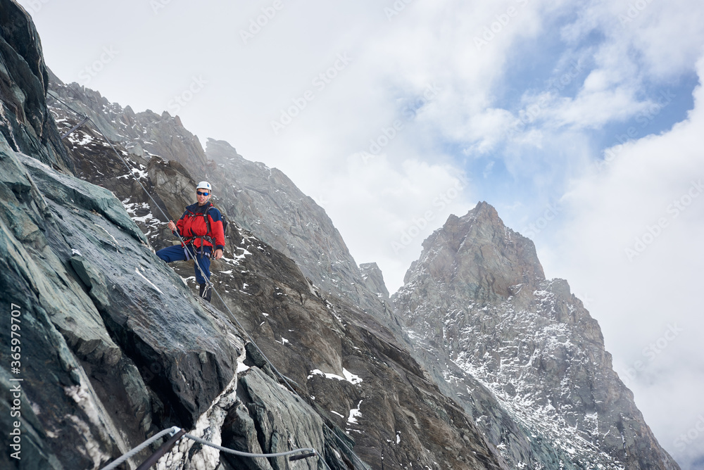 Young man in sunglasses and safety helmet holding fixed rope while climbing mountain. Alpinist ascending natural rock formation. Concept of mountaineering and alpine rock climbing.