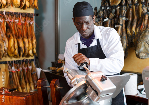 Confident African American seller slicing ham on slicer machine in butcher shop..