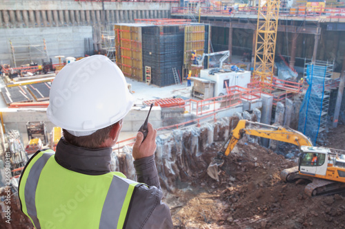 construction worker or civil engineer or architect or foreman with hard hat and safety vest at major construction site with crane and digger supervising with walkie talkie photo