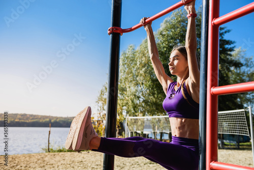 Portrait of strong young woman in sports clothing hanging on wall bars with her legs up. Fitness woman performing hanging leg raises on outdoor.